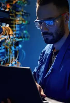 IT infrastructure technician holding a laptop while managing complex network cables in a server rack cabinet